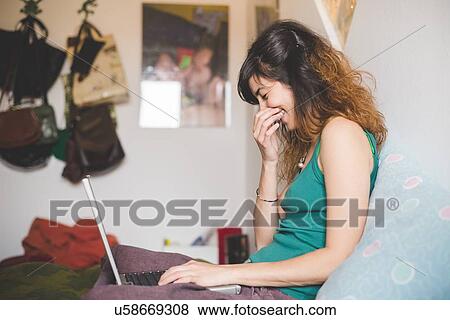 Side view of young woman sitting on bed, legs crossed, using laptop, hand to mouth, eyes closed laughing View Large Photo Image Stock Photo - Side view of young woman sitting on bed, legs crossed, using laptop, hand to mouth, eyes closed laughing. Fotosearch
