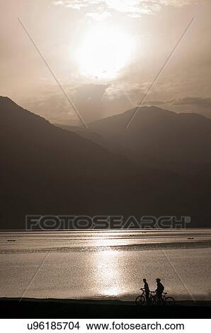 Picture - Silhouette of cyclists with bicycles viewing mountain and lake at sunset, Nepal. Fotosearch