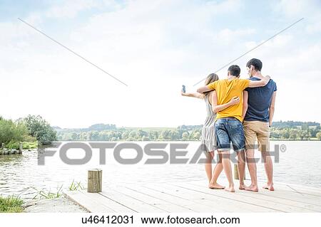Three young adults standing on jetty, taking self portrait, using smartphone, rear view View Large Photo Image Stock Image - Three young adults standing on jetty, taking self portrait, using smartphone, rear view. Fotosearch