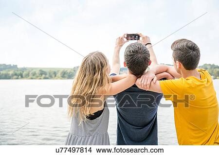 Three young adults taking self portrait using smartphone, rear view View Large Photo Image Picture - Three young adults taking self portrait using smartphone, rear view. Fotosearch