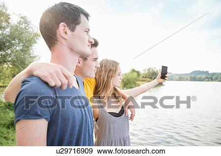 Stock Photo - Three young adults taking self portrait, using smartphone, outdoors. Fotosearch