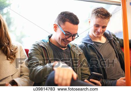 Stock Image - Two young male friends on train, looking at smartphone. Fotosearch