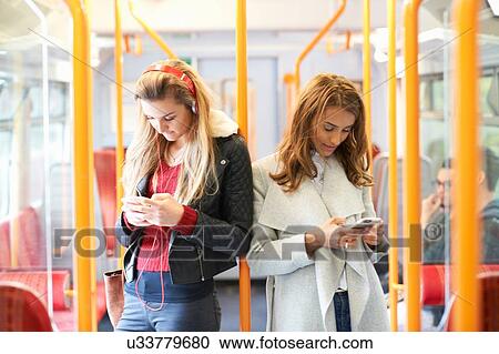 Stock Image - Two young women on train, back to back, using smartphones. Fotosearch