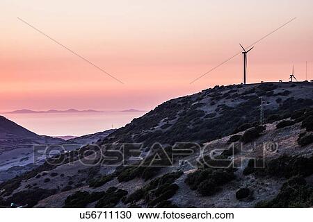 Stock Image - Wind turbines on mountain top against dramatic orange sky, Castelsardo, Sardinia, Italy. Fotosearch