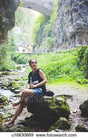 Stock Photo - Woman sitting on rock in stream, rock hills and stone bridge in background, Garda, Italy. Fotosearch