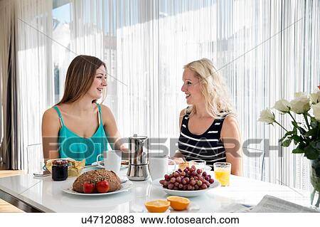 Stock Image - Women sitting at dining table enjoying a continental breakfast together, face to face smiling. Fotosearch