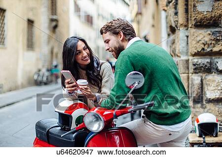 Stock Photo - Young couple leaning on moped reading smartphone texts, Florence, Italy. Fotosearch