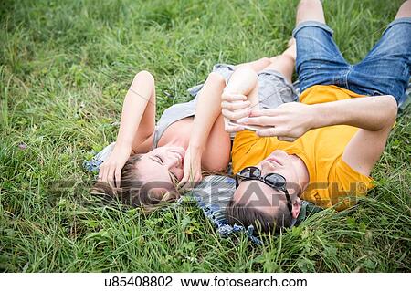 Stock Image - Young couple lying on grass in field, looking at smartphone. Fotosearch