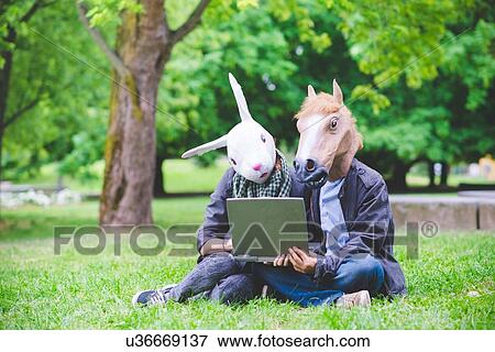 Young couple sitting on grass wearing rabbit and horse costume masks holding laptop computer View Large Photo Image Stock Photo - Young couple sitting on grass wearing rabbit and horse costume masks holding laptop computer. Fotosearch