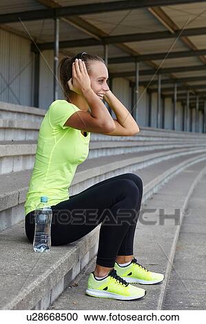 Stock Image - Young female runner adjusting headphones from stadium seating. Fotosearch