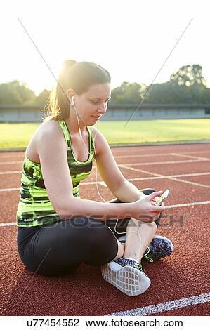 Stock Image - Young female runner cross legged on race track reading smartphone texts. Fotosearch