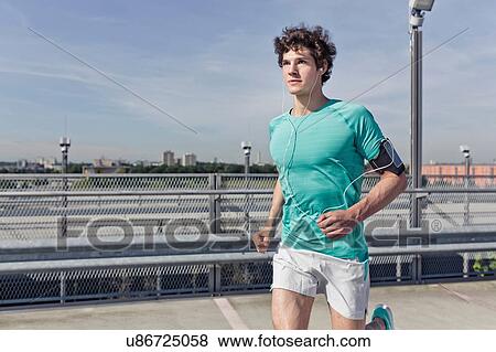 Young male runner wearing earphones whilst running in city View Large Photo Image Stock Photo - Young male runner wearing earphones whilst running in city. Fotosearch