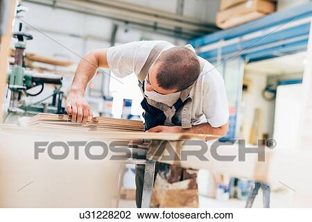 Stock Image - Young man in workshop looking down counting plywood sheets. Fotosearch