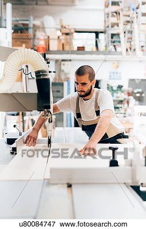 Stock Photo - Young man in workshop using carpentry machine to cut plywood. Fotosearch