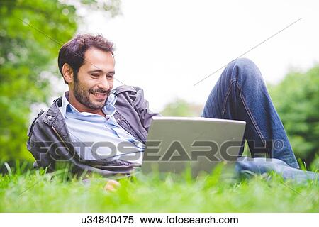 Stock Photography - Young man lying on grass using laptop computer looking down smiling. Fotosearch