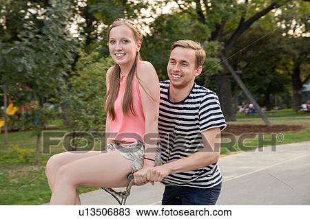 Stock Image - Young man riding bicycle with young woman sitting on handlebars smiling. Fotosearch