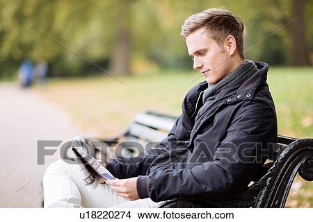 Young man sitting on bench reading newspaper in park View Large Photo Image Picture - Young man sitting on bench reading newspaper in park. Fotosearch