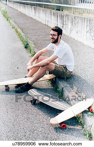 Stock Photograph - Young man sitting on kerb with three skateboards. Fotosearch
