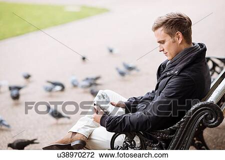 Young man sitting on park bench reading newspaper View Large Photo Image Stock Image - Young man sitting on park bench reading newspaper. Fotosearch