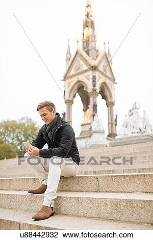 Stock Image - Young man sitting on steps reading smartphone texts, Albert Memorial, London, England, UK. Fotosearch