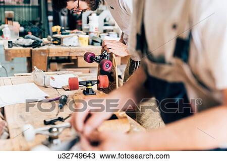 Stock Image - Young men in carpentry workshop looking down making skateboards. Fotosearch