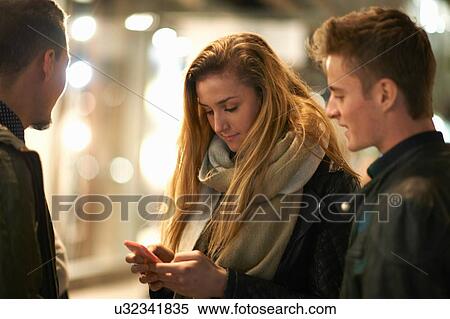 Stock Photography - Young woman and friends reading smartphone texts on street at night, London, UK. Fotosearch