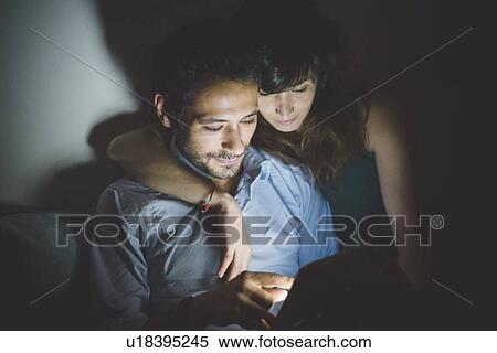 Stock Photography - Young woman sitting with arm around young man illuminated by laptop computer screen. Fotosearch