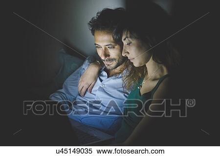 Stock Photography - Young woman sitting with arm around young man illuminated by laptop computer screen. Fotosearch