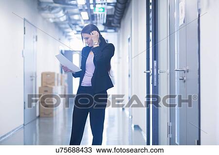 Stock Image - Young woman standing in office corridor looking down at paperwork scratching head. Fotosearch