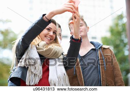 Stock Image - Young woman taking smartphone selfie with male friends. Fotosearch