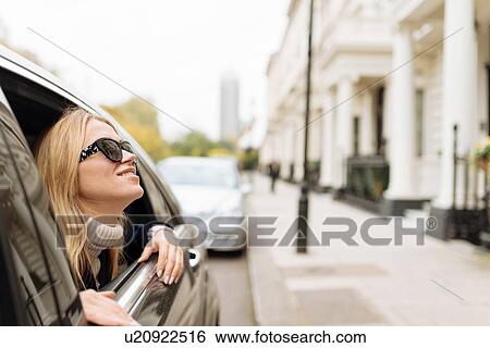 Stock Photograph - Young woman wearing sunglasses looking up from car window, London, England, UK. Fotosearch