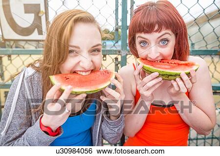Stock Photograph - Young women eating watermelon beside sports ground, London, UK. Fotosearch