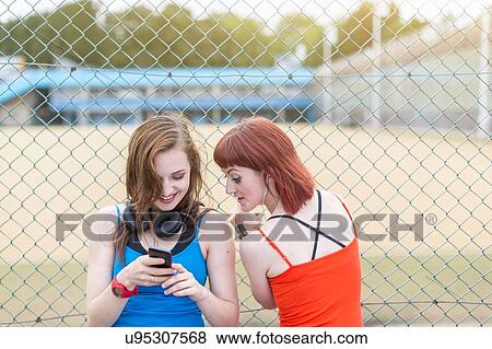Stock Photo - Young women using smartphone beside fence, London, UK. Fotosearch