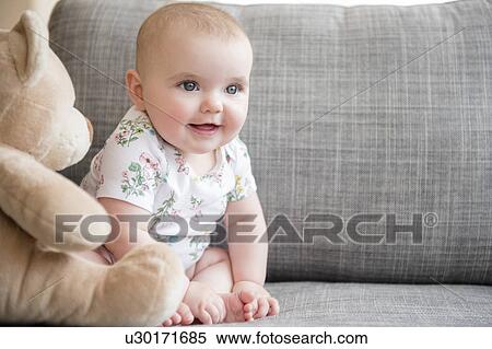 Stock Photography - Baby girl (12-17 months) with teddy bear sitting on sofa. Fotosearch