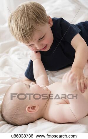 Stock Photograph - Brother (2-3) playing with baby sister (12-17 months) on bed. Fotosearch