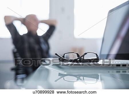 Stock Photo - Eyeglasses and laptop on desk in office, man relaxing in background. Fotosearch
