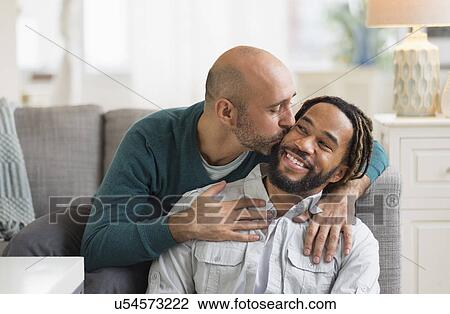 Stock Image - Homosexual couple kissing by sofa in living room. Fotosearch