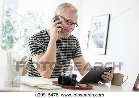 Stock Photograph - Man in home office using smartphone and tablet. Fotosearch