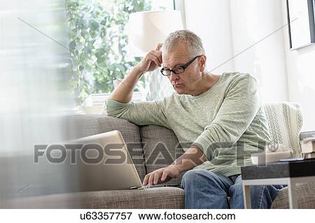 Man sitting on sofa using laptop at home View Large Photo Image Stock Photo - Man sitting on sofa using laptop at home. Fotosearch
