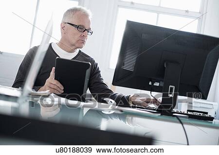 Stock Photo - Man working in office using computer and tablet. Fotosearch