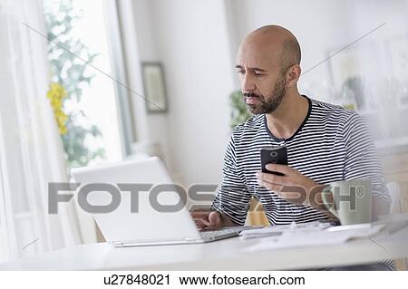 Stock Image - Man working with laptop and holding smart phone at table. Fotosearch