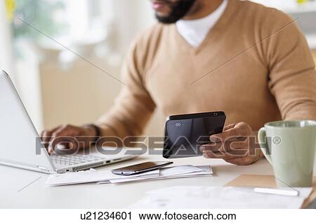 Stock Image - Man working with laptop at table and holding digital tablet. Fotosearch