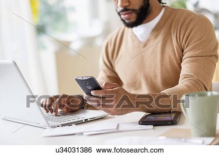 Stock Photograph - Man working with laptop at table and holding smart phone. Fotosearch