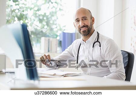 Stock Image - Portrait of smiley doctor working with laptop at desk in office. Fotosearch