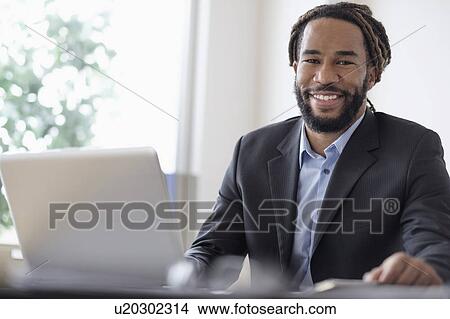 Picture - Smiley businessman sitting at desk with laptop. Fotosearch