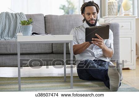 Picture - Young man with dreadlocks using digital table by sofa in living room. Fotosearch