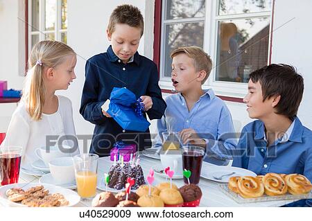 Stock Photo - Boy with siblings unwrapping birthday gifts on patio. Fotosearch