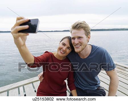 Couple on pier side by side using smartphone to take selfie smiling, Copenhagen, Denmark View Large Photo Image Stock Image - Couple on pier side by side using smartphone to take selfie smiling, Copenhagen, Denmark. Fotosearch