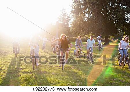 Stock Image - Crowds of adult friends arriving on bicycles to sunset park party. Fotosearch