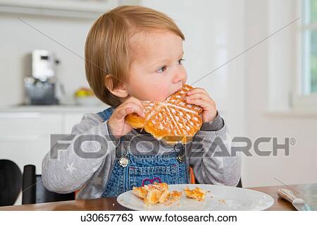Stock Image - Cute female toddler eating cake at kitchen table. Fotosearch
